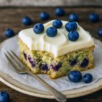 Close-up of a golden sheet cake studded with fresh blueberries and flecks of zucchini, topped with smooth lemon buttercream and a sprinkle of lemon zest.